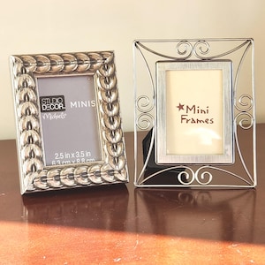 May include: Two silver mini picture frames. One frame has a textured, repeating design around the border. The other frame has a decorative, swirling design. The frames are sized 2.5 in x 3.5 in (6.3 cm x 8.8 cm). The text "Mini Frames" is visible.