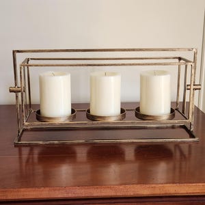 May include: A decorative candle holder with three ivory-colored pillar candles. The holder is made of gold-colored metal with a rectangular frame and individual circular trays for each candle. The candles are unscented.