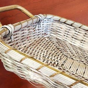 May include: A rectangular woven basket with a handle. The basket is made of light gray and tan woven material. The handle is a curved, tan-colored material. The basket is empty and sits on a brown surface.