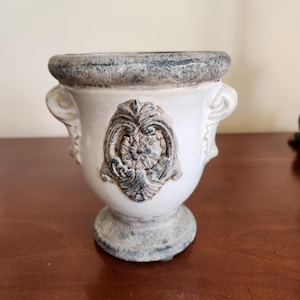 May include: A white ceramic urn-shaped planter featuring a weathered gray finish on the rim and base. The center of the vessel displays a raised, ornate floral relief design in a dark gray tone, complemented by two small side handles.