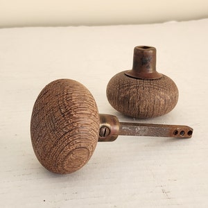 May include: Two antique wooden door knobs. One knob is oval-shaped, the other is round. Both knobs have a textured, wood grain appearance. The knobs are connected by a metal rod and are set against a white background.