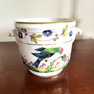 May include: A white porcelain cachepot featuring two side handles and a gold-trimmed rim. The exterior is decorated with a hand-painted scene of a green and blue bird perched on a branch, surrounded by small blue flowers and green foliage.