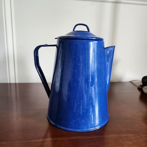 May include: A deep blue enamel coffee pot with a speckled white finish, featuring a tapered cylindrical body, a curved handle on the side, a pointed spout, and a matching removable lid with a small arched handle on top.