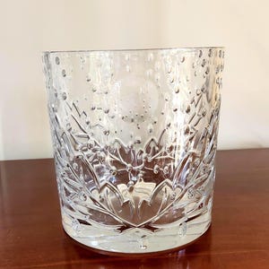 May include: A clear glass vessel featuring a cylindrical shape with a textured surface. The glass is adorned with a pattern of small, raised bubbles throughout the upper section and a series of sharp, angular leaf-like carvings at the base.