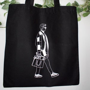 May include: Black tote bag with a white outline of a person wearing sunglasses and carrying a bag. The person is walking.