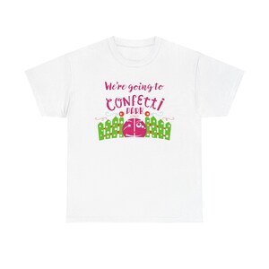 Puede incluir: Camiseta blanca con el texto "We're going to Confetti Park" en rosa y verde. El diseño incluye una valla verde, una bola rosa y blanca y elementos decorativos.