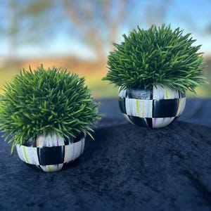 May include: Two small, round planters with vibrant green, spiky faux plants. The planters are painted with a black and white checkerboard pattern, accented with thin stripes of yellow, purple, and green. The planters sit on a dark blue surface.