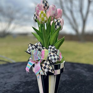May include: A decorative floral arrangement featuring pink and checkered tulips in a black and white striped pot. The arrangement is adorned with a checkered bow and colorful ribbons, creating a whimsical and stylish display.