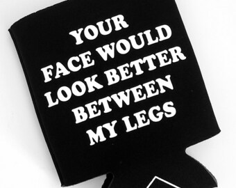 Op de afbeelding: Zwarte neopreen-blikjeskoeler met witte tekst die "YOUR FACE WOULD LOOK BETTER BETWEEN MY LEGS" luidt. De onderkant van de koeler heeft een wit logo met de tekst "KUNTX".