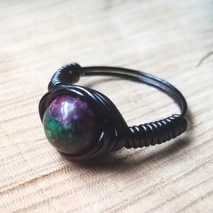 May include: A black wire-wrapped ring with a green, purple, and black gemstone.