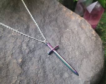 Puede incluir: Un collar de cadena plateada con un colgante de espada iridiscente arcoíris. El colgante está sobre una roca gris oscura con musgo verde en el fondo.