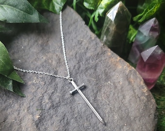 May include: A silver sword pendant necklace on a silver chain. The necklace is displayed on a gray rock with green foliage in the background.