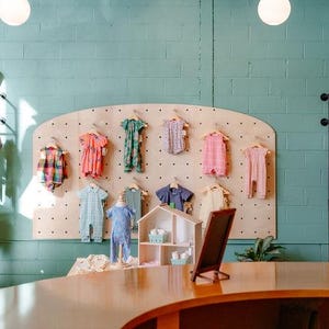 May include: A retail space featuring teal cinder block walls, light wood pegboard displays, and black metal clothing racks. Various children's garments hang on hangers, while a wooden dollhouse sits on a curved wooden counter in the foreground.