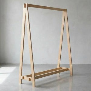 May include: A light-coloured wooden clothes rack with an A-frame design. The rack has a horizontal bar at the top for hanging clothes and a lower shelf for storage. The rack is made of light-coloured wood and stands on a concrete floor.