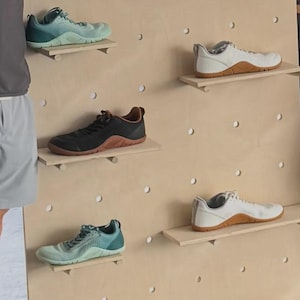 Op de afbeelding: Vijf sportschoenen tentoongesteld op een lichthouten pegboard-wand. De collectie bevat twee paar witte sneakers met gummizolen, twee paar groenblauwe sneakers met kleurverloop en &eacute;&eacute;n paar zwarte sneakers met een bruine gummizool, allemaal met veters.