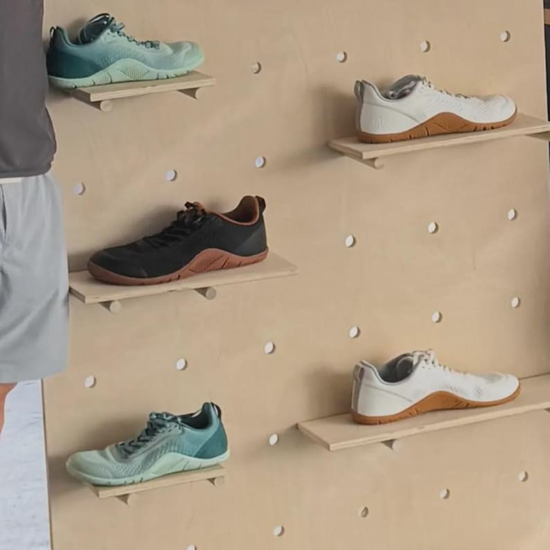 Op de afbeelding: Vijf sportschoenen tentoongesteld op een lichthouten pegboard-wand. De collectie bevat twee paar witte sneakers met gummizolen, twee paar groenblauwe sneakers met kleurverloop en &eacute;&eacute;n paar zwarte sneakers met een bruine gummizool, allemaal met veters.