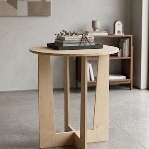May include: A light wood round side table with a unique leg design. The table has a black tray on top with books, greenery, and a compass. The table is approximately 24 inches in diameter and 20 inches tall.