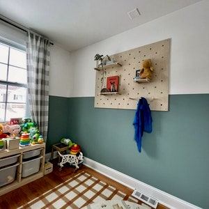 Puede incluir: Un tablero perforado rectangular grande de madera clara montado en una pared, con m&uacute;ltiples orificios circulares para estantes ajustables. El tablero sostiene estantes peque&ntilde;os de madera con libros, una planta peque&ntilde;a y un peluche.