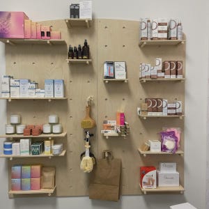 Op de afbeelding: Een lichtgekleurde houten pegboard-displaywand met meerdere horizontale planken met diverse huidverzorgingsproducten, verpakte cosmetica, kleine kaarsen, borstels en een bruine papieren tas die aan een haakje in het midden hangt.