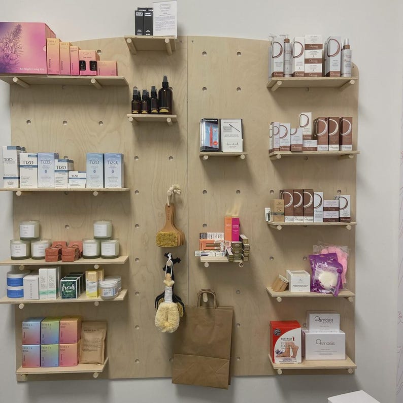 Op de afbeelding: Een lichtgekleurde houten pegboard-displaywand met meerdere horizontale planken met diverse huidverzorgingsproducten, verpakte cosmetica, kleine kaarsen, borstels en een bruine papieren tas die aan een haakje in het midden hangt.