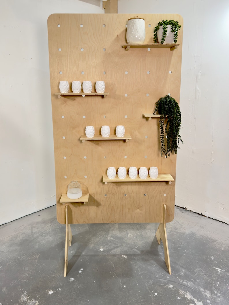 K&ouml;nnte beinhalten: Eine helle Holz-Lochwand-Ausstellung mit mehreren Regalen. Die Regale beherbergen wei&szlig;e Keramikvasen und dekorative Pflanzen. Die Lochwand hat eine Reihe von gleichm&auml;&szlig;ig verteilten L&ouml;chern und wird von Holzbeinen getragen.