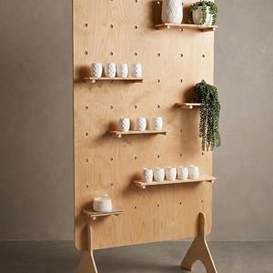 May include: A wooden display stand with several small shelves. The stand has a pegboard design and holds decorative items, including small white geometric patterned cups, potted plants, and a candle.