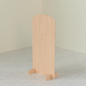 A light wood arched pegboard display stand with two support feet, viewed from a slight angle against a light-colored wall and patterned floor.