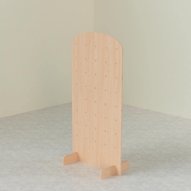 A light wood arched pegboard display stand with two support feet, viewed from a slight angle against a light-colored wall and patterned floor.