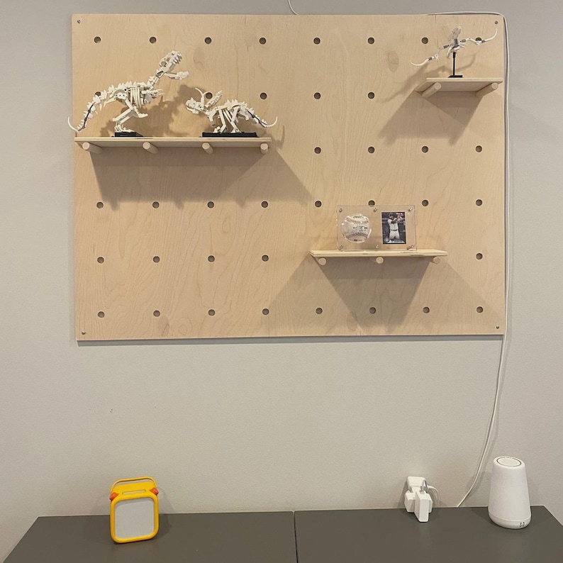 Op de afbeelding: Een lichtgekleurd houten pegboard wandorganizer met drie horizontale planken. Op de planken staan twee witte dinosaurus-skeletmodellen, een klein vliegend wezen, een honkbal in een doorzichtige houder en een kleine ingelijste foto.