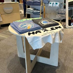 May include: A round wooden table with a white cloth and two stacks of t-shirts. The t-shirts have a blue and black color scheme with graphic designs and text that reads "Surf Down Belly" and "Adventure Waves." The table has a white cloth with the text "HA-LEN" printed on it.