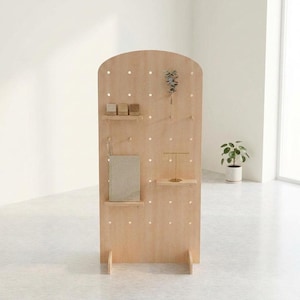 A light wood arched pegboard display stand with various items on its pegs, including small boxes, a book, and jewelry, in a minimalist room.