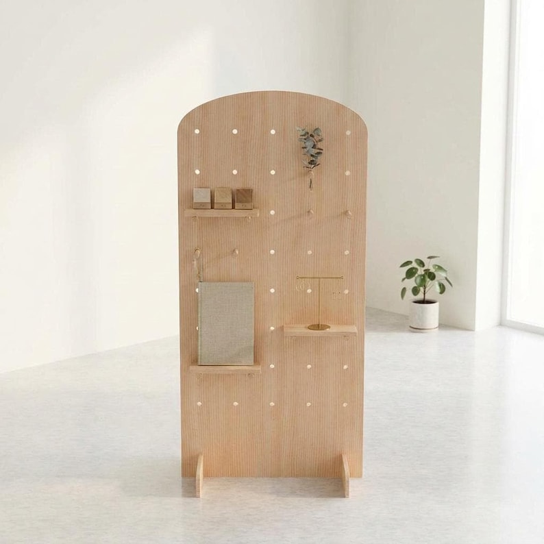 A light wood arched pegboard display stand with various items on its pegs, including small boxes, a book, and jewelry, in a minimalist room.