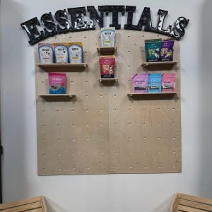 Op de afbeelding: Een lichtgekleurde houten pegboard displaywand met het woord ESSENTIALS in grote zwarte letters bovenaan. Verschillende kleine houten planken zijn aan het bord bevestigd en bevatten diverse kleurrijke zakjes met dierensnacks van merken zoals Arch.