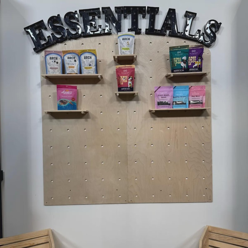 Op de afbeelding: Een lichtgekleurde houten pegboard displaywand met het woord ESSENTIALS in grote zwarte letters bovenaan. Verschillende kleine houten planken zijn aan het bord bevestigd en bevatten diverse kleurrijke zakjes met dierensnacks van merken zoals Arch.