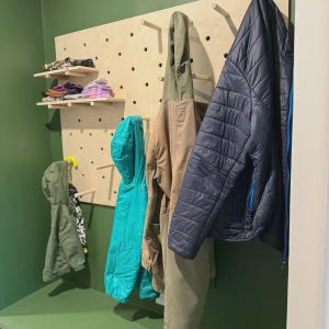 Puede incluir: Un panel perforado de madera clara para pared con m&uacute;ltiples clavijas cil&iacute;ndricas para colgar objetos. La unidad incluye dos estantes peque&ntilde;os con ropa doblada y varias chaquetas azules, beige y verdes colgadas de las clavijas.