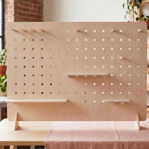 A large, light wood pegboard display stand with two shelves and several pegs, set on a table with a pink cloth in a bright room.