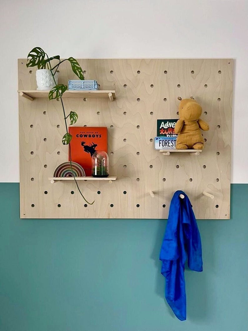 Puede incluir: Un tablero perforado rectangular grande de madera clara con una cuadr&iacute;cula de agujeros circulares. El tablero incluye dos estantes horizontales que sostienen una planta en maceta, un libro titulado The Book of Cowboys, un arco&iacute;ris y un peluche.