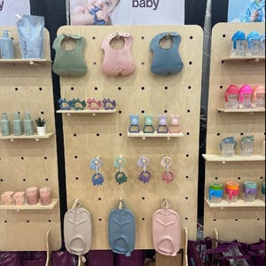 K&ouml;nnte beinhalten: Ausstellung von Babyprodukten, darunter L&auml;tzchen, Bei&szlig;ringe und Trinkbecher, die auf Holz-Lochw&auml;nden angeordnet sind. Die L&auml;tzchen sind in Gr&uuml;n-, Rosa- und Blaut&ouml;nen gehalten. Die Ausstellung zeigt auch Babyflaschen und Beh&auml;lter. Das Wort "baby" ist sichtbar.
