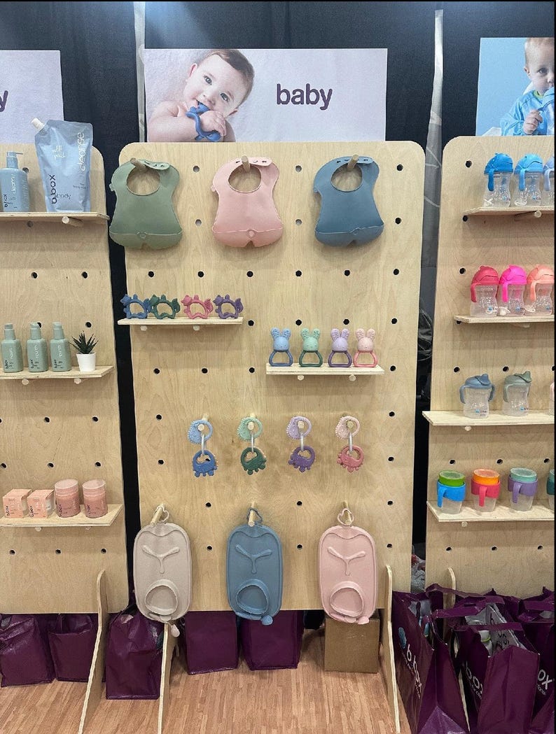 K&ouml;nnte beinhalten: Ausstellung von Babyprodukten, darunter L&auml;tzchen, Bei&szlig;ringe und Trinkbecher, die auf Holz-Lochw&auml;nden angeordnet sind. Die L&auml;tzchen sind in Gr&uuml;n-, Rosa- und Blaut&ouml;nen gehalten. Die Ausstellung zeigt auch Babyflaschen und Beh&auml;lter. Das Wort "baby" ist sichtbar.