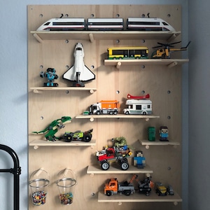Op de afbeelding: Een lichtgekleurde houten pegboard wanddisplay met meerdere verstelbare planken waarop diverse plastic bouwsteenmodellen staan, waaronder een witte trein, een spaceshuttle, een gele bus, een groene dinosaurus en twee doorzichtige emmers met kleine steentjes.