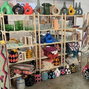 May include: A wooden shelf unit filled with colorful ceramic vases, woven baskets, and patterned mugs. The shelves are decorated with a variety of items, including rugs, blankets, and other home decor.