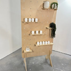 K&ouml;nnte beinhalten: Ein h&ouml;lzerner Ausstellungsst&auml;nder mit Lochwand-Design und mehreren Regalen. Er zeigt wei&szlig;e Keramikvasen und ein dekoratives Gef&auml;&szlig; sowie gr&uuml;ne Pflanzen in wei&szlig;en T&ouml;pfen. Der St&auml;nder ist aus hellem Holz gefertigt und dient zur Pr&auml;sentation von Artikeln.