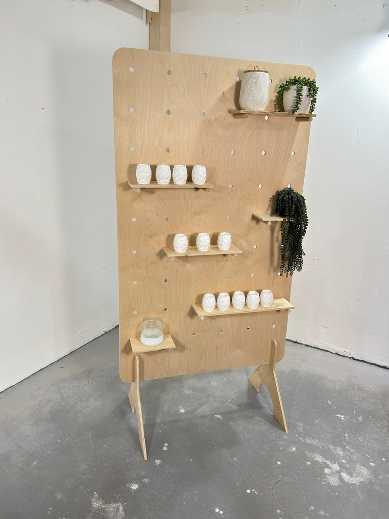 K&ouml;nnte beinhalten: Ein h&ouml;lzerner Ausstellungsst&auml;nder mit Lochwand-Design und mehreren Regalen. Er zeigt wei&szlig;e Keramikvasen und ein dekoratives Gef&auml;&szlig; sowie gr&uuml;ne Pflanzen in wei&szlig;en T&ouml;pfen. Der St&auml;nder ist aus hellem Holz gefertigt und dient zur Pr&auml;sentation von Artikeln.
