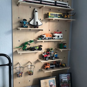 Op de afbeelding: Een grote, lichtgekleurde houten pegboard-wanddisplay met meerdere horizontale planken waarop diverse voertuigen van bouwstenen, een spaceshuttle, een dinosaurus en twee metalen emmers gevuld met kleine kleurrijke bouwsteentjes staan.