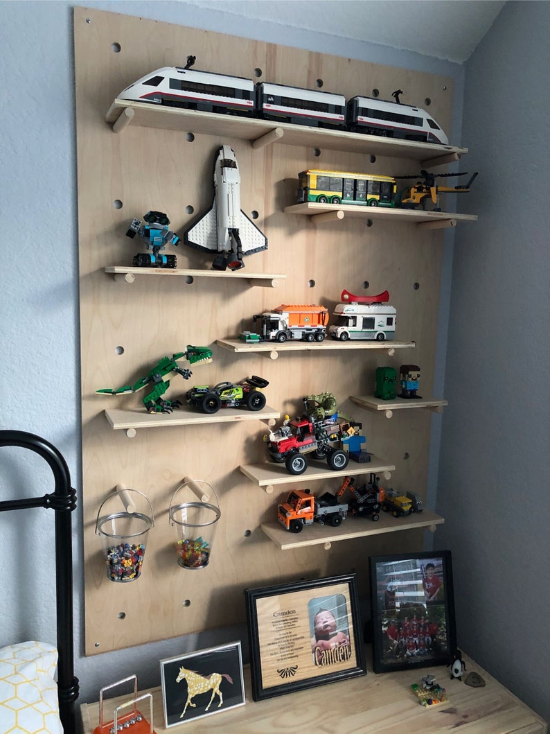 Op de afbeelding: Een grote, lichtgekleurde houten pegboard-wanddisplay met meerdere horizontale planken waarop diverse voertuigen van bouwstenen, een spaceshuttle, een dinosaurus en twee metalen emmers gevuld met kleine kleurrijke bouwsteentjes staan.
