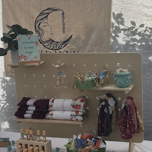 A light wood pegboard display stand with various textile products, small bottles, and a ceramic pot, set against a fabric banner with a moon design.