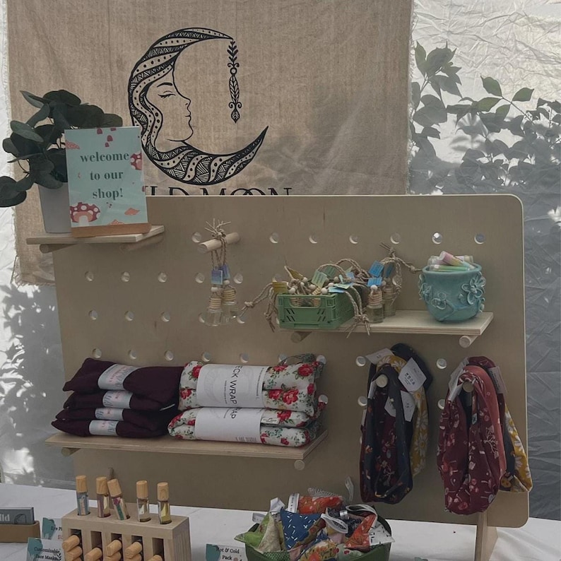 A light wood pegboard display stand with various textile products, small bottles, and a ceramic pot, set against a fabric banner with a moon design.