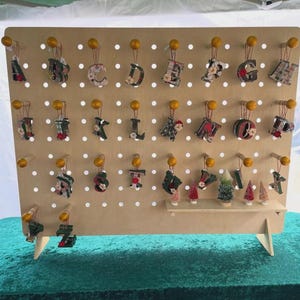 A light wood pegboard display stand with small, fabric-covered letter ornaments hanging from pegs, and miniature Christmas trees on shelves.