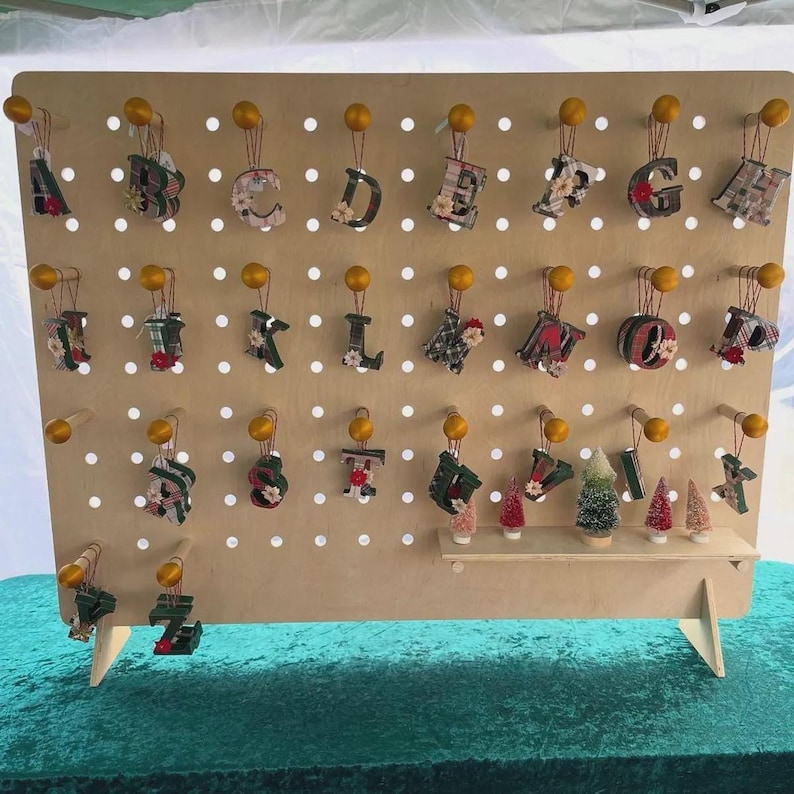 A light wood pegboard display stand with small, fabric-covered letter ornaments hanging from pegs, and miniature Christmas trees on shelves.