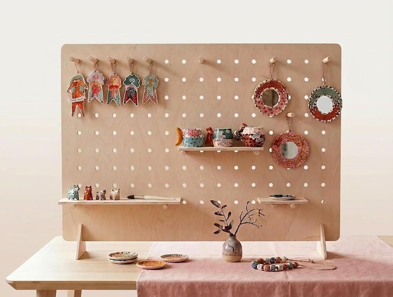 A light wood pegboard display stand with various colorful ceramic items and small figurines on shelves and pegs, set on a light pink tablecloth.